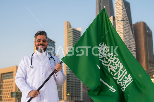 تقديم الرعاية الصحة لابناء الوطن، الاحتفالات بذكرى اليوم الوطني السعودي 23 سبتمبر، صورة مقربة لطبيب عربي خليجي سعودي يرتدي المعطف الأبيض يمسك بيده علم السعودية بإيماءات الفخر والاعتزاز, يوم العلم 11 مارس، خلفية أبراج مدينة الرياض وضح النهار