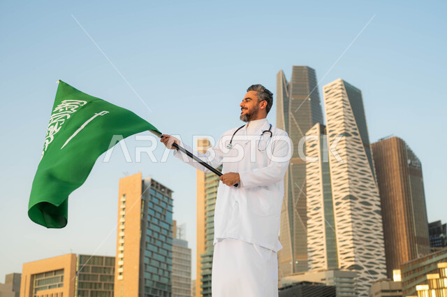 الاحتفالات بذكرى اليوم الوطني السعودي 23 سبتمبر، طبيب عربي خليجي سعودي يرتدي المعطف الأبيض يمسك بيده علم السعودية بإيماءات الفخر والاعتزاز, يوم العلم 11 مارس، تقديم الرعاية الصحة لابناء الوطن، خلفية مركز الملك عبدالله المالي (كافد) في مدينة الرياض