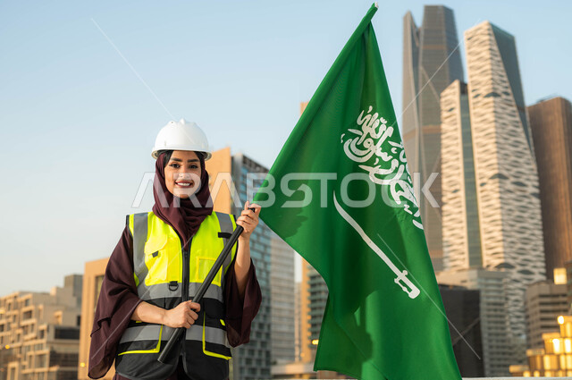 ايماءات السعادة بالاحتفال في يوم العلم 11مارس, دور المهندسات السعوديات في التطور العمراني، ذكرى اليوم الوطني السعودي 23 سبتمبر, صورة مقربة لمهندسة عربية سعودية خليجية محجبة ترتدي سترة وخوذة الحماية تحمل علم السعودية وضح النهار, خلفية مركز الملك عبدلله المالي