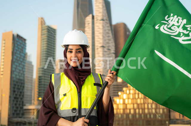 دور المهندسات السعوديات في التطور العمراني, ايماءات السعادة بالاحتفال في يوم العلم 11مارس,   ذكرى اليوم الوطني السعودي 23 سبتمبر, صورة مقربة لمهندسة عربية سعودية خليجية محجبة ترتدي سترة وخوذة الحماية تحمل علم السعودية وضح النهار, خلفية أبراج مدينة الرياض