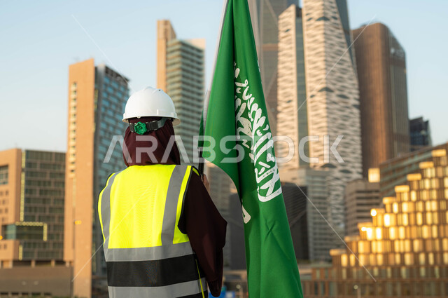 Participation of the Kingdom’s women in its urban development, commemoration of the Saudi National Day, September 23, a picture from behind of a veiled Saudi Arabian Gulf engineer wearing a helmet and protection vest, holding the Kingdom’s flag in broad daylight, celebrating Flag Day, March 11, background of the towers of the city of Riyadh