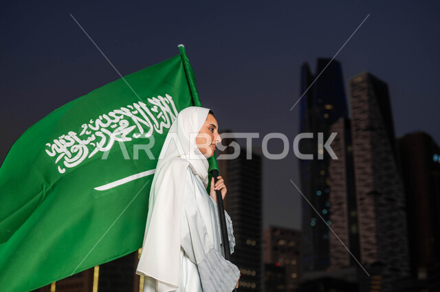 ايماءات الفخر والتباهي بامتلاك الهوية السعودية, تخليد يوم العلم 11 مارس, الاحتفال بذكرى اليوم الوطني السعودي 23 سبتمبر, صورة من الجانب لامرأة عربية سعودية خليجية مبتمسة ترتدي العباءة والحجاب تمسك علم المملكة في وقت الليل, خلفية أبراج مدينة الرياض 