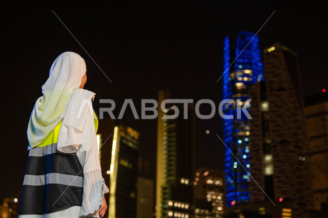 الاهتمام بالتطوير العمراني, صورة من الخلف لمهندسة عربية خليجية سعودية ترتدي سترة الحماية والحجاب تنظر الى مركز الملك عبدالله المالي(كافد) في وقت الليل, المعالم السياحية المشهورة في السعودية, خلفية الأبراج المالية والمنشآت الاقتصادية في مدينة الرياض 