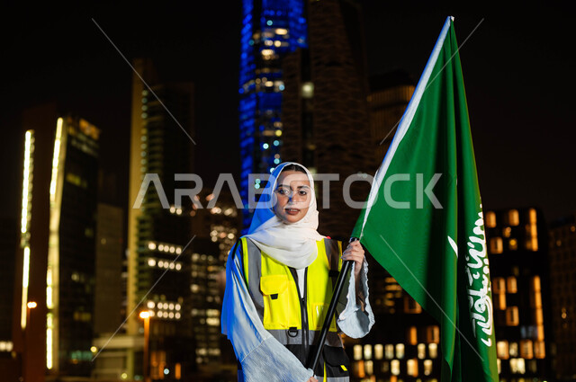 مشاركة نساء المملكة بتطورها العمراني, إحياء ذكرى اليوم الوطني السعودي 23 سبتمبر, مهندسة عربية سعودية خليجية ترتدي الحجاب وسترة الحماية تمسك علم المملكة وقت الليل، النظر الى الكاميرا بايماءات الفخر والاعتزاز، الاحتفال بيوم العلم 11 مارس, خلفية أبراج الرياض 