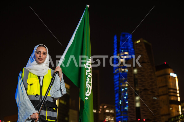 مهن ووظائف نسائية، إبراز دور النساء في التطور المعماري, إحياء ذكرى اليوم الوطني 23 سبتمبر, صورة مقربة لمهندسة عربية سعودية خليجية مبتمسة ترتدي الحجاب وسترة الحماية تمسك علم المملكة تنظر الى الكاميرا, يوم العلم 11 مارس, خلفية أبراج مدينة الرياض وقت الليل