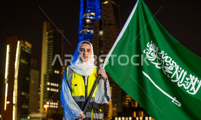 مهن ووظائف نسائية، إبراز دور النساء في التطور المعماري, إحياء ذكرى اليوم الوطني 23 سبتمبر, صورة مقربة لمهندسة عربية سعودية خليجية مبتمسة ترتدي الحجاب وسترة الحماية تمسك علم المملكة تنظر الى الكاميرا, يوم العلم 11 مارس, خلفية أبراج مدينة الرياض وقت الليل