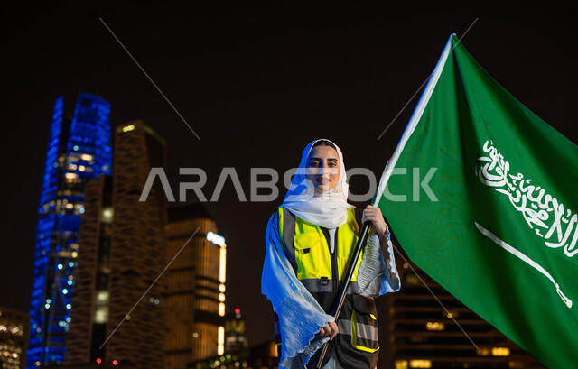 يوم العلم 11 مارس, مهن ووظائف نسائية، إبراز دور النساء في التطور المعماري, إحياء ذكرى اليوم الوطني 23 سبتمبر, مهندسة عربية سعودية خليجية مبتمسة ترتدي الحجاب وسترة الحماية تمسك علم المملكة تنظر الى الكاميرا, خلفية أبراج مدينة الرياض وقت الليل