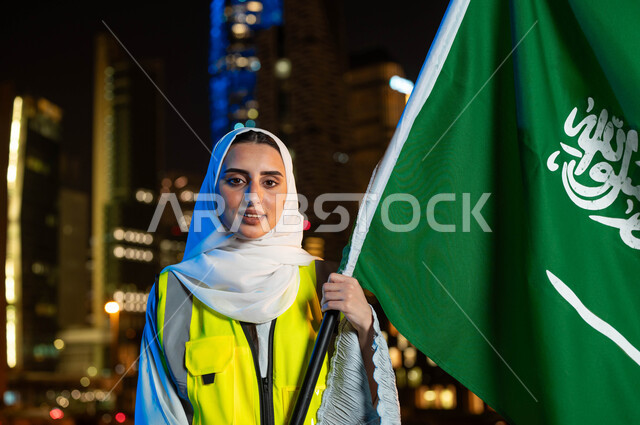 إبراز دور النساء في التطور المعماري, مهن ووظائف نسائية، إحياء ذكرى اليوم الوطني 23 سبتمبر, صورة مقربة لمهندسة عربية سعودية خليجية مبتمسة ترتدي الحجاب وسترة الحماية تمسك علم المملكة تنظر الى الكاميرا, يوم العلم 11 مارس, خلفية أبراج مدينة الرياض وقت الليل