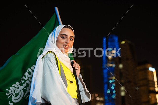 إحياء ذكرى اليوم الوطني السعودي, دور المهندسات السعوديات في التطور العمراني, الاحتفال بتوحيد المملكة في 8 ربيع الأول, صورة مقربة لمهندسة عربية سعودية خليجية محجبة ترتدي سترة الحماية تمسك علم السعودية بيدها, يوم العلم 11مارس, خلفية أبراج مدينة الرياض وقت الليل