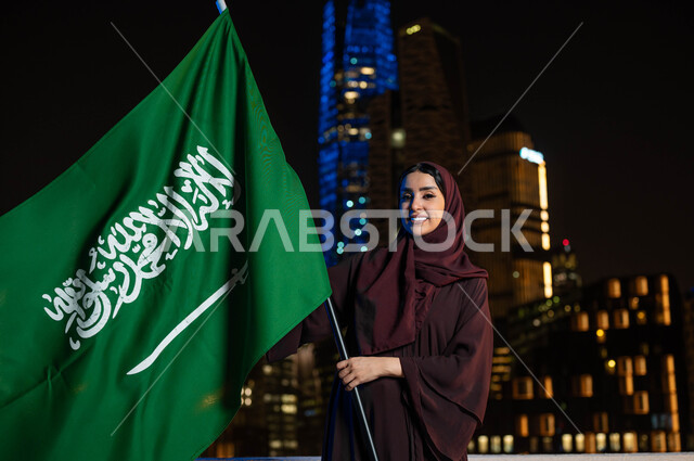 اليوم الوطني 8 ربيع الأول, الفخر و الاعتزاز بالانتماء للمملكة, ذكرى العيد الوطني السعودي, امرأة عربية سعودية خليجية ترتدي العباءة والحجاب تحمل بيدها علم السعودية للاحتفال بيوم العلم 11مارس, خلفية أبراج مدينة الرياض المضيئة ليلا