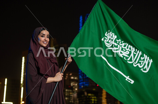 ذكرى العيد الوطني السعودي, الفخر و الاعتزاز بالانتماء للمملكة, اليوم الوطني 8 ربيع الأول, امرأة عربية سعودية خليجية ترتدي العباءة والحجاب تحمل بيدها علم السعودية للاحتفال بيوم العلم 11مارس, خلفية أبراج مدينة الرياض المضيئة ليلا