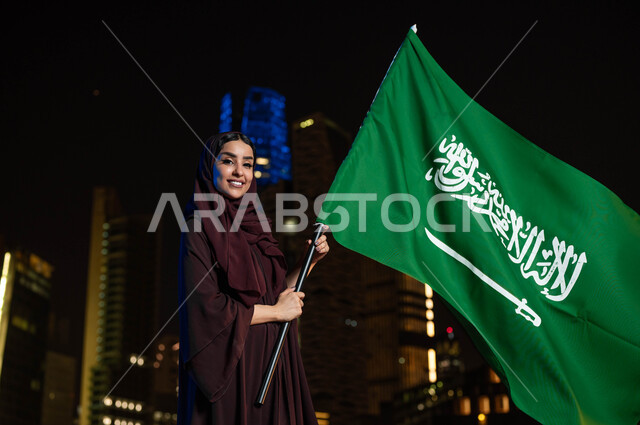 الفخر و الاعتزاز بالتطور العمراني للمملكة, ذكرى العيد الوطني السعودي, اليوم الوطني 8 ربيع الأول, امرأة عربية سعودية خليجية ترتدي العباءة والحجاب تحمل بيدها علم السعودية للاحتفال بيوم العلم 11مارس, خلفية أبراج مدينة الرياض المضيئة ليلا