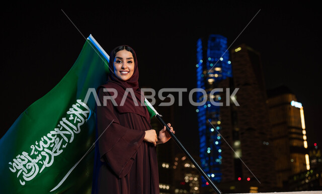 العيد الوطني في 23سبتمبر, الاحتفال بذكرى اليوم الوطني السعودي, امرأة عربية سعودية خليجية ترتدي العباءة والحجاب و تحمل بيدها علم المملكة بإيماءات الفخر, يوم العلم  11 مارس, خلفية أبراج مدينة الرياض ليلا