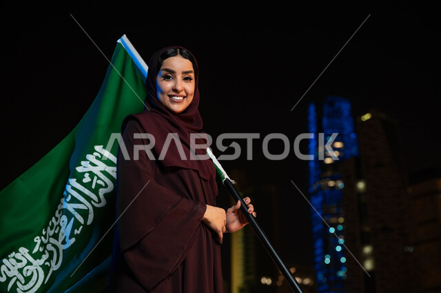 Saudi Flag Day, March 11, celebration of the National Day, a smiling Saudi Gulf Arab woman wearing the abaya and hijab and holding the Kingdom’s flag in her hand, National Day on September 23, the background of the illuminated towers of Riyadh at night time