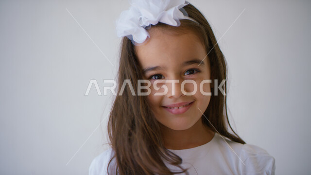 ارتداء الملابس الجديده في الأعياد والمناسبات، التقاط صور شخصية للوثائق المدرسية, الإهتمام بالمظهر الخارجي للأطفال الصغار، بورتريه مقرب لطفلة عربيه خليجيه إماراتية تنظر إلى الكاميرا مبتسمة ترتدي فستان أبيض اللون، خلفيه بيضاء