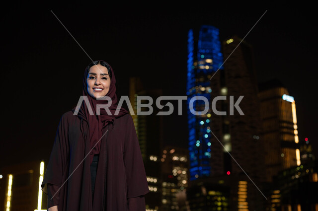 الاعتزاز والفخر بالتقدم المعماري في السعودية، النمو والتطور العمراني في مدينة الرياض، التجول والتنزه واستنشاق الهواء النقي في الخارج، إمرأة عربية خليجية سعودية مبتسمة ترتدي الحجاب تنظر الى الكاميرا، خلفية الابراج وناطحات السحاب المضيئة ليلا