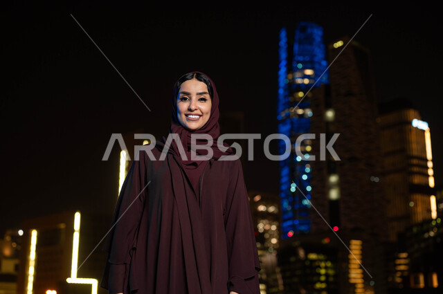 Pride and pride in architectural progress in Saudi Arabia, urban growth and development in the city of Riyadh, walking around and breathing fresh air outside, a smiling Saudi Gulf Arab woman wearing a hijab looking at the camera, the background of the towers and skyscrapers illuminated at night