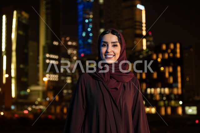 التطور والنمو العمراني في مدينة الرياض، الفخر والاعتزاز بالتقدم المعماري في السعودية، التجول والتنزه واستنشاق الهواء النقي في الخارج، صورة مقربة لإمرأة عربية خليجية سعودية مبتسمة ترتدي الحجاب تنظر الى الكاميرا، خلفية الابراج وناطحات السحاب المضيئة ليلا
