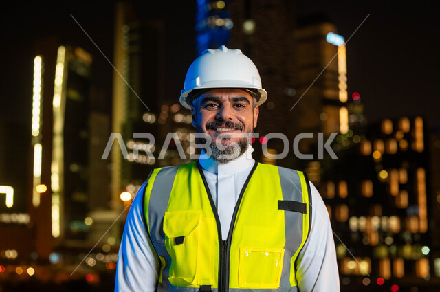 النظر إلى الكاميرا بايماءات السعاده، مفهوم الهندسة المعمارية والانشاءات, اماكن ومعالم سياحيه مشهورة ،النمو والتطور العمراني بسواعد أبناء الوطن, صورة مقربة لمهندس عربي خليجي سعودي يرتدي سترة العمل وخوذة الحماية البيضاء، خلفية  الأبراج المضاءة ليلا في الرياض .