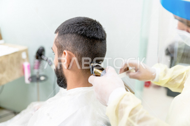 A Saudi Arabian Gulf man visits a barber shop. The barber uses the razor to tidy up the customer’s hair, taking care of the appearance.