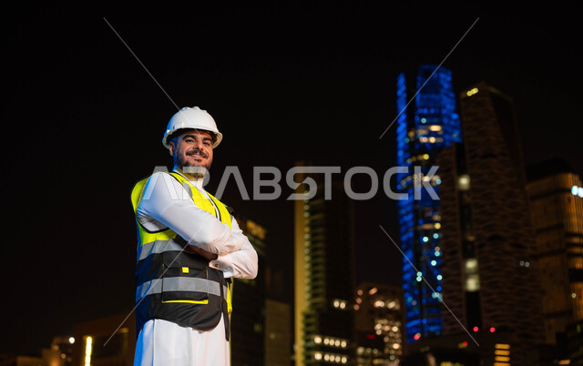 مفهوم الهندسة المعمارية والانشاءات, الوقوف بإيماءات أيدي متقاطعه، اماكن ومعالم سياحيه مشهورة ،النمو والتطور العمراني بسواعد أبناء الوطن, مهندس عربي خليجي سعودي يرتدي سترة العمل وخوذة الحماية البيضاء، خلفية ناطحات السحاب و الأبراج المضاءة ليلا في الرياض .