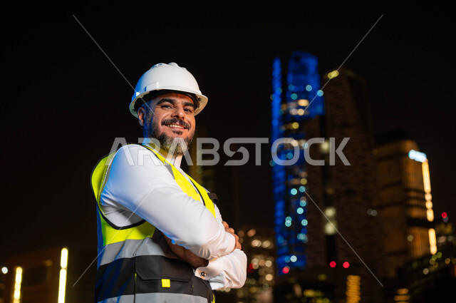 الوقوف بإيماءات أيدي متقاطعه، مفهوم الإنشاءات و الهندسة المعمارية, اماكن ومعالم سياحيه مشهورة ،النمو والتطور العمراني بسواعد أبناء الوطن, صورة مقربة لمهندس عربي خليجي سعودي يرتدي سترة العمل وخوذة الحماية البيضاء، خلفية الأبراج المضاءة ليلا في الرياض .