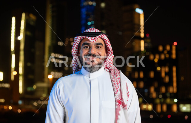 معالم سياحيه مشهورة ، التقدم والتطور العمراني في المدينة، صورة مقربة لرجل عربي خليجي سعودي مبتسم يرتدي الشماغ و الزي السعودي التقليدي ينظر الى الكاميرا، مفهوم الاهتمام بالمظهر الخارجي، خلفية الأبراج وناطحات السحاب المضيئة ليلا في مدينة الرياض