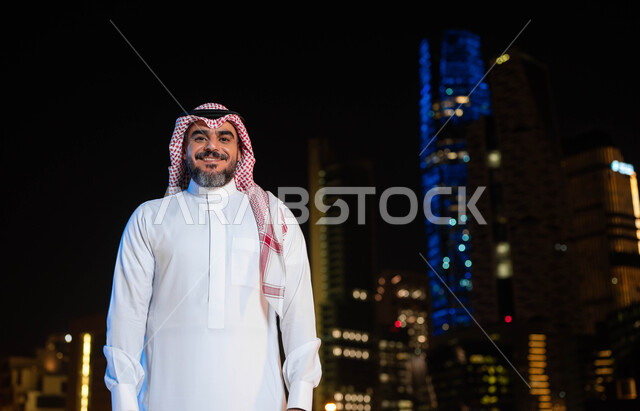 التقدم والتطور العمراني في المدينة، معالم سياحيه مشهورة ،رجل عربي خليجي سعودي مبتسم يرتدي الشماغ و الزي السعودي التقليدي ينظر الى الكاميرا، مفهوم الاهتمام بالمظهر الخارجي، خلفية الأبراج وناطحات السحاب المضيئة ليلا في مدينة الرياض