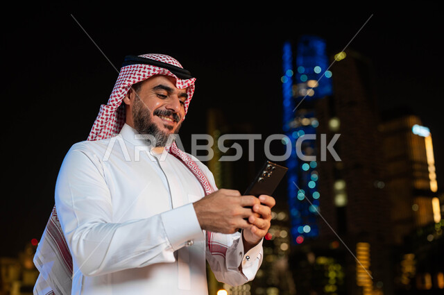 استخدام أجهزة تقنية حديثة، الدردشة والتواصل مع الأصدقاء عبر برامج التواصل الاجتماعي، صورة مقربة لرجل عربي خليجي سعودي مبتسم يرتدي الشماغ و الزي التقليدي يحمل بيده الهاتف المحمول، خلفية الأبراج وناطحات السحاب المضيئة ليلا في مدينة الرياض