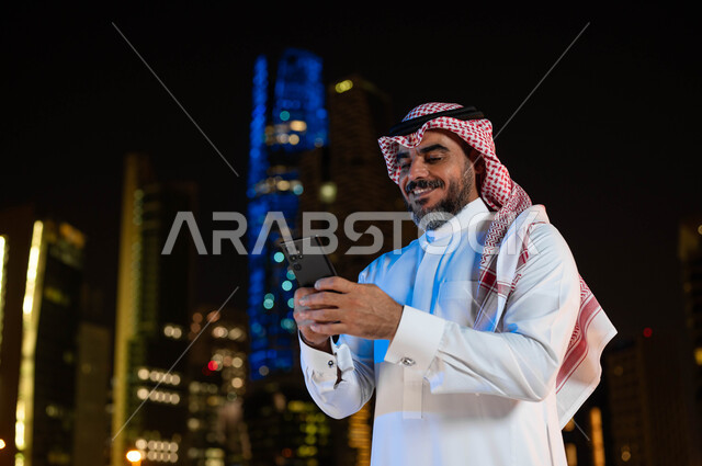 الدردشة والتواصل مع الأصدقاء عبر برامج التواصل الاجتماعي، استخدام أجهزة تقنية حديثة، رجل عربي خليجي سعودي مبتسم يرتدي الشماغ و الزي التقليدي يحمل بيده الهاتف المحمول، خلفية الأبراج وناطحات السحاب في مدينة الرياض ليلا، معالم سياحيه مشهورة