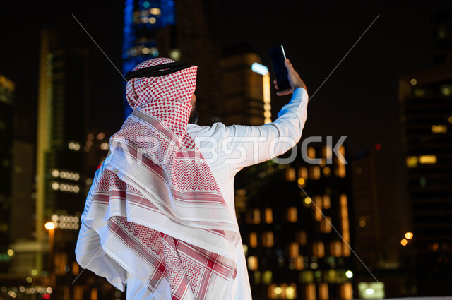 الوقوف الأبراج وناطحات السحاب المضيئة ليلا في مدينة الرياض، التقدم والتطور العمراني في المدينة، صورة من الخلف لرجل عربي خليجي سعودي يرتدي الشماغ و الزي التقليدي يقوم بالتقاط صور سيلفي تذكارية باستخدام الهاتف المحمول, معالم سياحيه مشهورة