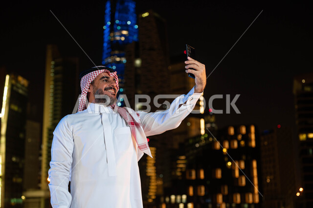 Chat and communicate with friends and make video calls, a smiling Saudi Gulf Arab man wearing a shemagh and traditional dress taking souvenir selfies using a mobile phone, the background of the illuminated towers and skyscrapers at night in Riyadh, famous tourist attractions