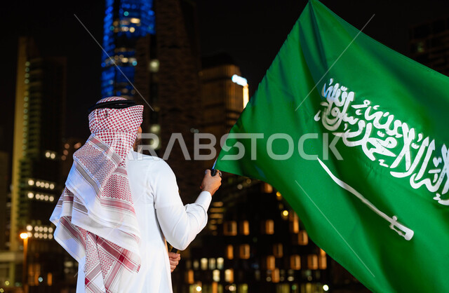 مناسبات و احتفالات وطنية شعبية، اظهار الولاء والاخلاص للبلاد ذكرى اليوم الوطني السعودي 23 سبتمبر، صورة من الخلف لرجل عربي خليجي سعودي يرتدي الشماغ والثوب في العيد الوطني يحمل بيده علم المملكة، يوم العلم 11 مارس، الوقوف أمام الابراج المضيئة ليلا في الرياض