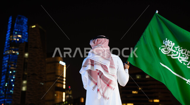 احتفالات ومناسبات وطنية شعبية، اظهار الولاء والاخلاص للبلاد ذكرى اليوم الوطني السعودي 23 سبتمبر، صورة من الخلف لرجل عربي خليجي سعودي يرتدي الشماغ والثوب في العيد الوطني يحمل بيده علم المملكة، يوم العلم 11 مارس، الوقوف أمام الابراج المضيئة ليلا في الرياض