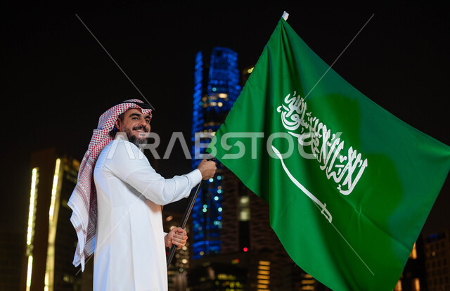 Showing loyalty and love for the country, commemorating the Saudi National Day, Rabi’ al-Awwal 8, a smiling Saudi Gulf Arab man wearing a thobe and shemagh on the National Day holding the Kingdom’s flag in his hand, celebrating Flag Day, March 11, national events and holidays, the background of the towers and illuminated skyscrapers at night in Riyadh.