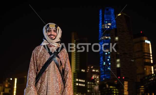The popular traditional costume of the central region, celebrations of national occasions, the anniversary of the Saudi National Day September 23, the day of the founding of the first Saudi state in 1727 AD, a smiling Saudi Arab man wearing the Dakla and Ghutra on the National Day looking at the camera, the background of illuminated skyscrapers at night