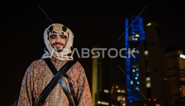 الزي الشعبي للمنطقة الوسطى، الاحتفالات بالمناسبات الوطنية، ذكرى اليوم الوطني السعودي 23 سبتمبر، يوم تأسيس الدولة السعودية الاولى 1727 م، صورة مقربة لرجل عربي سعودي مبتسم يرتدي الدقلة والغترة في العيد الوطني ينظر إلى الكاميرا، خلفية الابراج المضيئة ليلا في الرياض