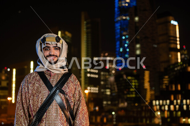 الزي الشعبي للمنطقة الوسطى، الاحتفالات بالمناسبات الوطنية، ذكرى اليوم الوطني السعودي 23 سبتمبر، يوم تأسيس الدولة السعودية الاولى 1727 م، صورة مقربة لرجل عربي سعودي مبتسم يرتدي الدقلة والغترة في العيد الوطني ينظر إلى الكاميرا، خلفية ناطحات السحاب المضيئة ليلا