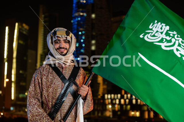اليوم الوطني السعودي 23 سبتمبر، ذكرى يوم التأسيس في 22 فبراير ،الأزياء الشعبيه بالمناسبات الوطنية، يوم العلم 11 مارس، صورة مقربة لرجل عربي خليجي سعودي مبتسم يرتدي الدقلة والغترة في العيد الوطني يحمل بيده علم المملكة، خلفية الابراج المضيئة ليلا في الرياض