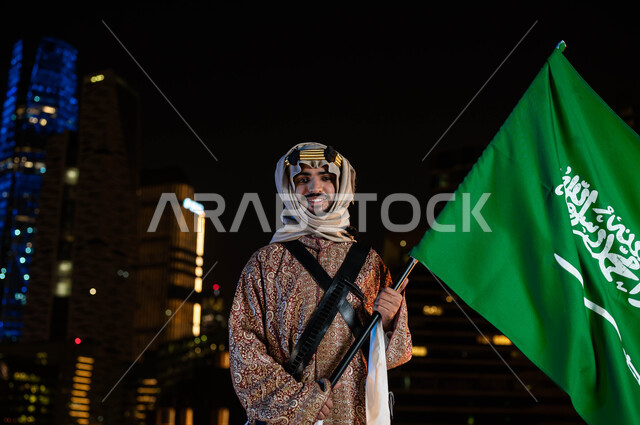 ذكرى يوم التأسيس في 22 فبراير ،الأزياء الشعبيه للاحتفالات بالمناسبات الوطنية، يوم العلم 11 مارس، اليوم الوطني السعودي 23 سبتمبر، رجل عربي خليجي سعودي مبتسم يرتدي الدقلة والغترة في العيد الوطني يحمل بيده علم المملكة، خلفية ناطحات السحاب المضيئة ليلا في الرياض
