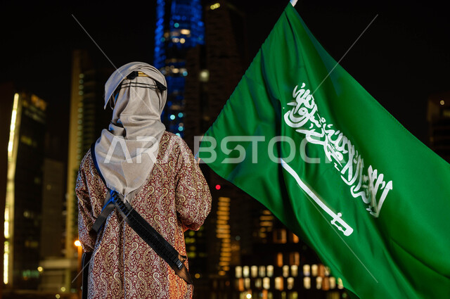 تأسيس الدولة السعوية الاولى 1727 م، يوم العلم 11 مارس، أزياء الاحتفالات بالمناسبات الوطنية، ذكرى اليوم الوطني السعودي 23 سبتمبر، صورة من الخلف لرجل خليجي سعودي يرتدي الدقلة والغترة في العيد الوطني يحمل بيده علم المملكة، يقف أمام أبراج الرياض المضيئة ليلا