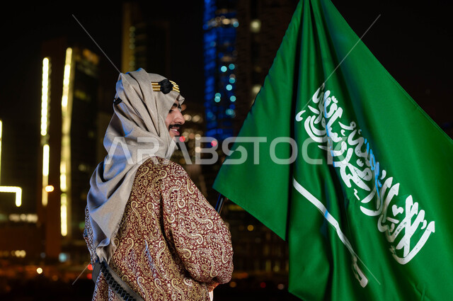 الاحتفالات بالمناسبات الوطنية، يوم العلم ١١ مارس، تأسيس الدولة السعودية 1727 م، ذكرى اليوم الوطني السعودي 23 سبتمبر، صورة من الخلف لرجل عربي خليجي سعودي مبتسم يرتدي الدقلة والغترة في العيد الوطني يحمل بيده علم المملكة، الوقوف أمام الابراج المضيئة ليلا في الرياض