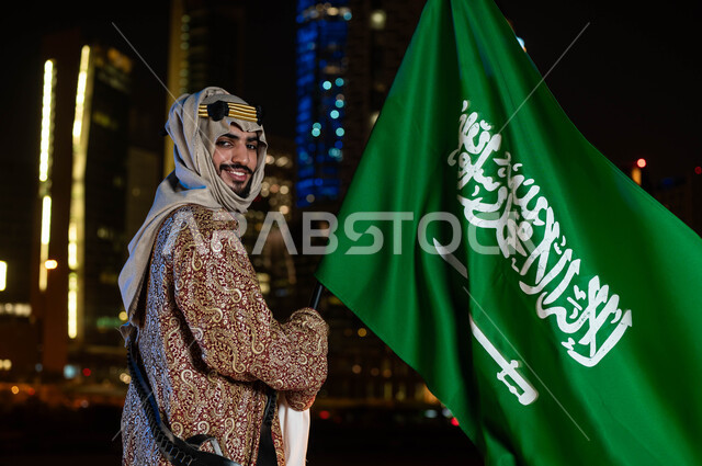 الاحتفالات بالمناسبات و الاعياد الوطنية، يوم العلم ١١ مارس، الزي الشعبي التقليدي للمنطقة الوسطى، ذكرى اليوم الوطني السعودي 23 سبتمبر، رجل خليجي سعودي مبتسم يرتدي الدقلة والغترة يحمل بيده علم المملكة، خلفية التطور العمراني في المباني والابراج في الرياض