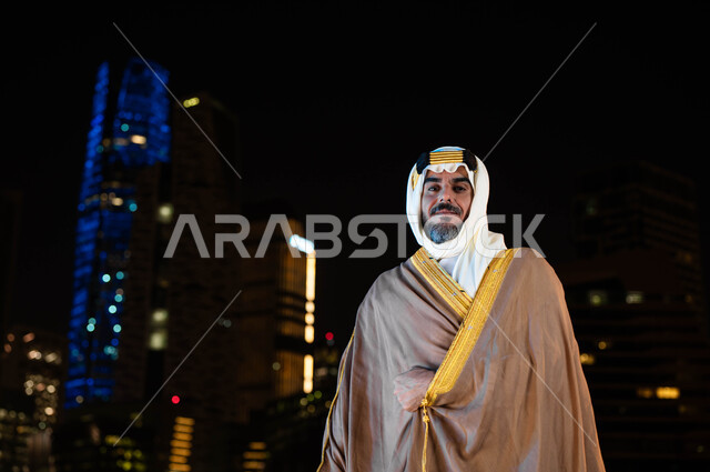استذكار التاريخ وتعزيز الهوية الوطنية، اظهار الحب والولاء للبلاد، تأسيس الدولة السعودية 1727 م، ذكرى اليوم الوطني السعودي 23 سبتمبر، رجل عربي خليجي سعودي مبتسم يرتدي البشت والغترة في العيد الوطني و ينظر إلى الكاميرا، خلفية ناطحات السحاب المضيئة ليلا في الرياض