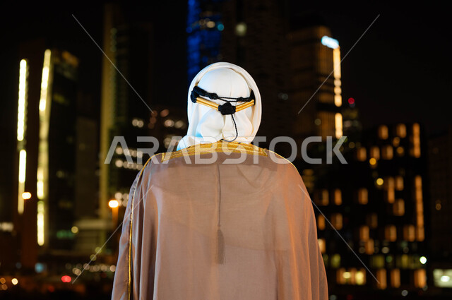 Expressing loyalty and love for the country, celebrating the anniversary of the Saudi National Day, September 23, the founding of the Saudi state in 1727 AD, a close-up photo from the back of a Saudi Gulf Arab man wearing a bisht and ghutra on National Day, standing in front of the towers and illuminated skyscrapers at night in the city of Riyadh.