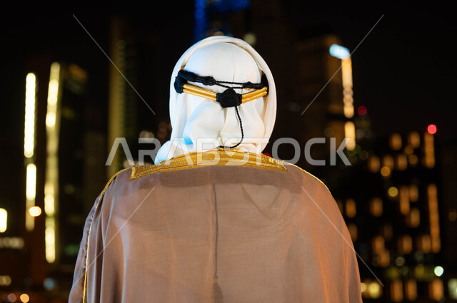 Expressing loyalty and love for the country, celebrating the anniversary of the Saudi National Day, September 23, the founding of the Saudi state in 1727 AD, a close-up photo from the back of a Saudi Gulf Arab man wearing a bisht and ghutra on National Day, standing in front of the towers and illuminated skyscrapers at night in the city of Riyadh.