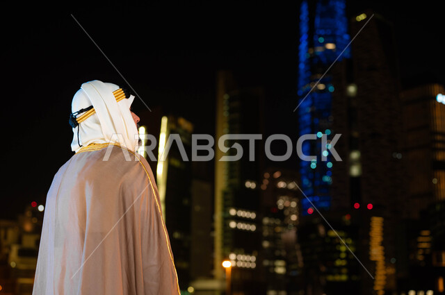 التعبير عن الحب والولاء للبلاد، الاحتفال بذكرى اليوم الوطني السعودي 23 سبتمبر، تأسيس الدولة السعودية 1727 م، صورة من الخلف لرجل عربي خليجي سعودي يرتدي البشت والغترة في العيد الوطني يقف أمام الابراج وناطحات السحاب المضيئة ليلا في مدينة الرياض