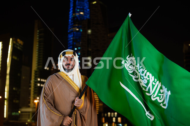 اظهار التفاني والانتماء للبلاد، تأسيس الدولة السعودية 1727 م، يوم العلم 11 مارس، الاحتفال بذكرى اليوم الوطني السعودي 23 سبتمبر، رجل عربي خليجي سعودي مبتسم يرتدي البشت والغترة في العيد الوطني يحمل بيده علم المملكة، خلفية ناطحات السحاب المضيئة ليلا في الرياض