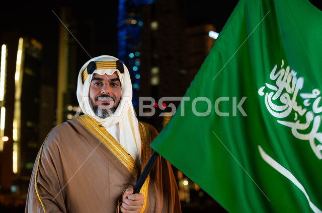 استذكار الهوية الوطنية والتاريخ، تأسيس الدولة السعودية 1727 م، يوم العلم 11 مارس، ذكرى اليوم الوطني السعودي 23 سبتمبر، صورة مقربة لرجل عربي خليجي سعودي مبتسم يرتدي البشت والغترة في العيد الوطني يحمل بيده علم المملكة، خلفية ناطحات السحاب المضيئة ليلا في الرياض