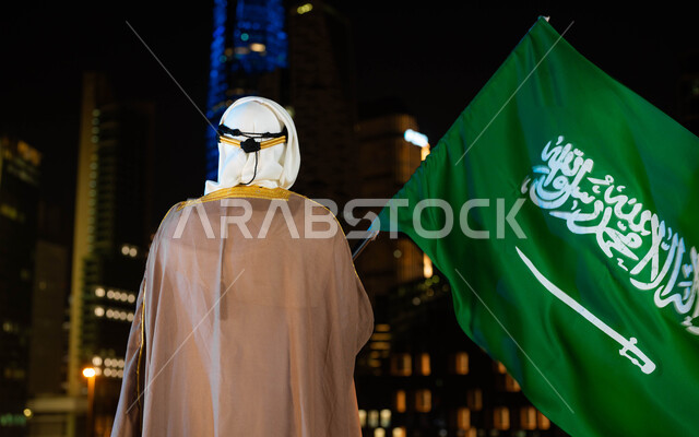 الاحتفال بذكرى اليوم الوطني السعودي 23 سبتمبر، تأسيس الدولة السعودية 1727 م، يوم العلم 11 مارس، صورة من الخلف لرجل عربي خليجي سعودي يرتدي البشت والغترة في العيد الوطني يحمل بيده علم المملكة، الوقوف أمام الابراج وناطحات السحاب المضيئة ليلا في مدينة الرياض